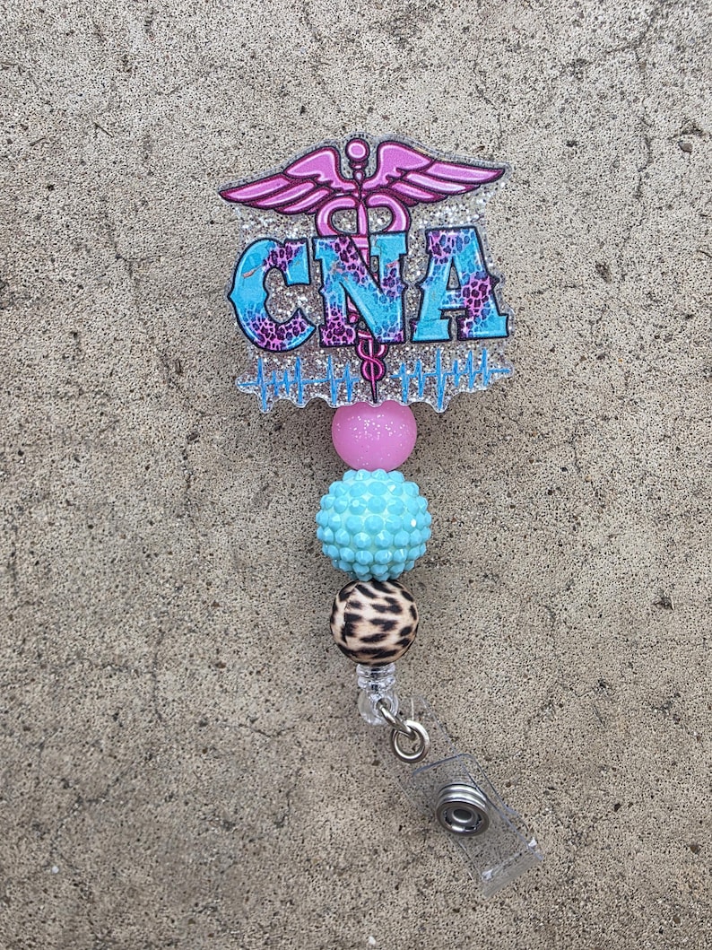 May include: A badge reel featuring a glittery acrylic topper with a pink caduceus symbol, the text CNA in blue and purple leopard print, and a blue heartbeat line. Below the topper are three beads: one pink, one textured light blue, and one leopard print.