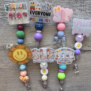 May include: Four badge reels featuring acrylic toppers with speech therapy themes. Designs include text like Squad, Everyone Communicates Differently, SLP, and Speech Therapy. Each reel has a vertical strand of colourful, textured, and patterned beads.