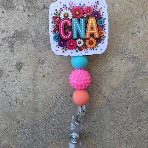 May include: A badge reel featuring a square topper with the text CNA in colorful, polka-dotted letters surrounded by a vibrant floral print. Below the topper are three beads: one smooth turquoise, one textured hot pink, and one smooth peach-colored bead.