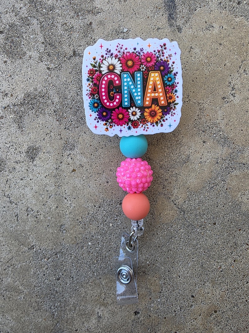 May include: A badge reel featuring a square topper with the text CNA in colorful, polka-dotted letters surrounded by a vibrant floral print. Below the topper are three beads: one smooth turquoise, one textured hot pink, and one smooth peach-colored bead.