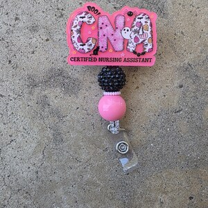 May include: A pink badge reel featuring the letters CNA in a Halloween-themed pattern with ghosts, pumpkins, and spiders. Below the letters, the text reads CERTIFIED NURSING ASSISTANT. The reel includes a black textured bead and a smooth pink bead.