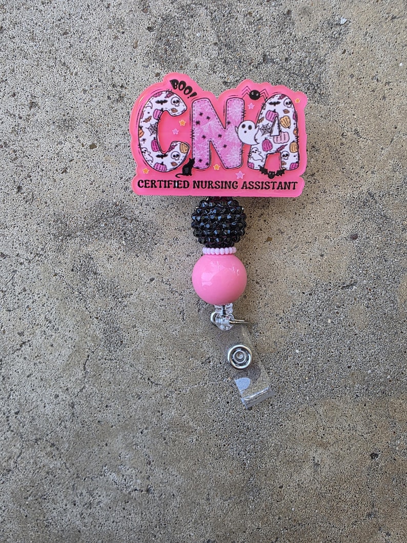 May include: A pink badge reel featuring the letters CNA in a Halloween-themed pattern with ghosts, pumpkins, and spiders. Below the letters, the text reads CERTIFIED NURSING ASSISTANT. The reel includes a black textured bead and a smooth pink bead.