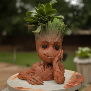 May include: A brown Groot-shaped planter with a green plant sprouting from the top. The planter has a detailed wood-like texture and black eyes. It is resting on a round, light-colored surface.