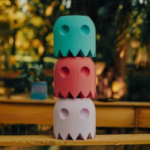 Puede incluir: Una pila de tres figuras de madera con forma de fantasma y colores. La figura superior es turquesa, la del medio coral y la inferior lavanda. Cada una tiene agujeros circulares para los ojos y un borde inferior dentado.