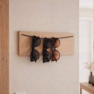 Puede incluir: Un soporte de madera para gafas de sol con tres pares de gafas colgando de un alambre negro. Las gafas de sol son negras y marrones. El soporte está montado en una pared de color claro.