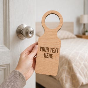 May include: A light-coloured wooden door hanger with a circular handle and a rectangular body. The text "YOUR TEXT HERE" is engraved on the front. The door hanger is held near a white door with a silver doorknob.