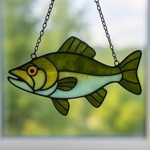 Puede incluir: Atrapasoles de pez de vidriera en tonos verdes y azul claro, con un contorno y cadena de metal negro para colgar. El pez tiene un ojo naranja y está suspendido sobre un fondo verde borroso.