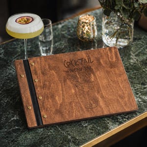 May include: A wooden cocktail menu with the words "COCKTAIL MENU BAR" engraved on the front. The menu is brown with a black spine and brass rivets. A cocktail glass with a passion fruit garnish sits next to the menu.