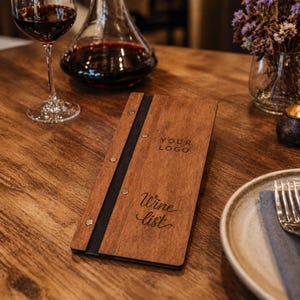 Könnte beinhalten: Ein hölzernes Weinlistenmenü mit den Worten "Your Logo" und "Wine List" auf der Vorderseite eingraviert. Das Menü ist braun mit einem schwarzen Rücken und Messingnieten. Ein Glas Rotwein und eine Karaffe befinden sich im Hintergrund.