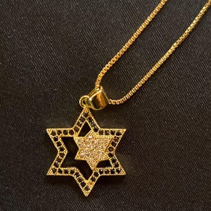 May include: Gold-toned Star of David pendant necklace. The pendant features a six-pointed star with a central star design. The outer star is outlined with black stones, and the inner star is filled with clear stones. The necklace chain is also gold-toned.