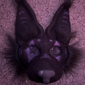 May include: A black furry mask with purple accents and mesh eyes. The mask has a pointed nose and large ears.