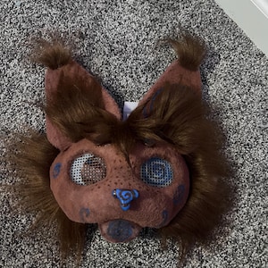 May include: A brown furry mask with blue and white details. The mask has a large nose and two large eyes with mesh coverings. The mask is designed to look like an animal.
