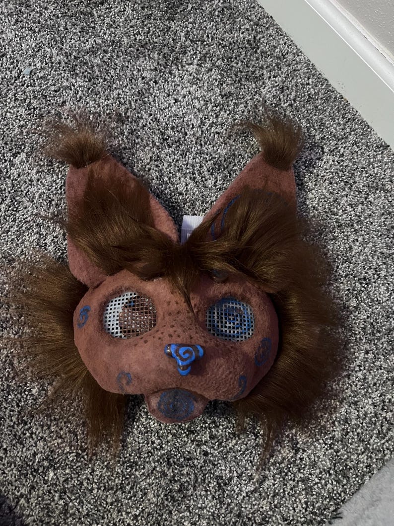 May include: A brown furry mask with blue and white details. The mask has a large nose and two large eyes with mesh coverings. The mask is designed to look like an animal.