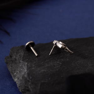 May include: Two silver-colored stud earrings. One earring has a small, round, dark-colored top. The other earring has a unique, geometric design. Both earrings have a post for insertion. The earrings are displayed on a dark, textured surface.