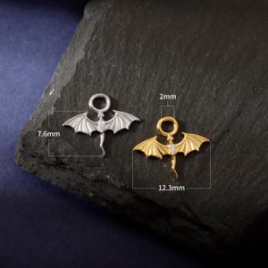 May include: Two dragon-shaped charms, one silver and one gold, each with a small gemstone. The silver charm is 7.6mm long, the gold charm 12.3mm. Both have a 2mm ring at the top for attachment. Ideal for jewellery making.