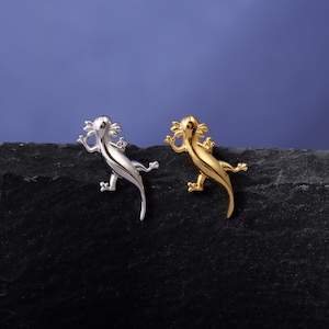 Puede incluir: Dos broches con forma de lagarto, uno plateado y otro dorado, se exhiben sobre una superficie oscura y texturizada contra un fondo azul. Los broches están detallados con patas, cola y cabeza con flecos, mostrando un diseño único.