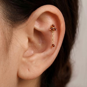 May include: A gold cartilage earring featuring a triangular cluster of three small amber-colored stones at the top, connected by a delicate gold chain to a single matching amber-colored stone dangling at the bottom, secured in the ear.