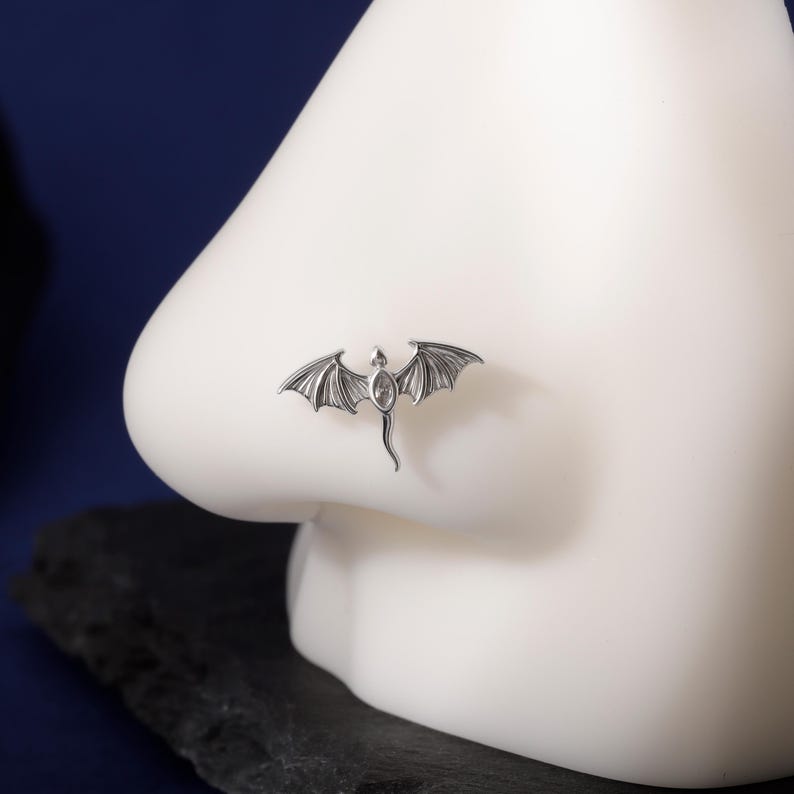 May include: A silver-colored nose ring in the shape of a dragon with outstretched wings. The dragon has a small, oval-shaped gem in its center. The nose ring is displayed on a white mannequin nose against a dark blue background.