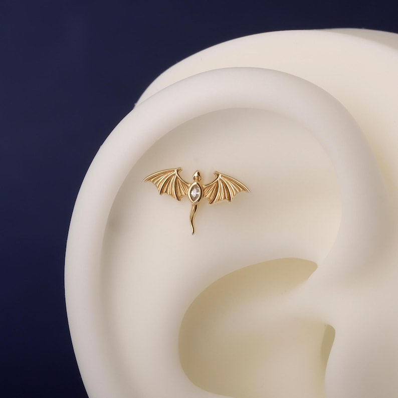 May include: A gold-colored dragon-shaped earring with a clear gemstone in the center. The dragon has detailed wings and a slender tail. The earring is displayed on a white ear model against a dark blue background.