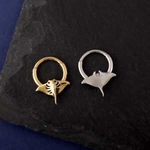 May include: Two hoop earrings with a manta ray design. One earring is gold-colored, and the other is silver-colored. The manta ray pendants are attached to the bottom of the hoops. The earrings are displayed on a dark, textured surface.