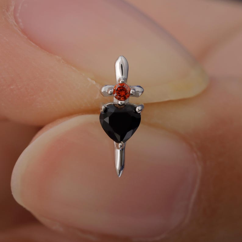 May include: A silver-colored earring with a black heart-shaped gem at the bottom, a small red gem in the middle, and a cross-shaped design at the top. The earring has a dagger-like shape.