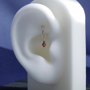 May include: A silver earring featuring a small, red heart-shaped gemstone pendant. The earring hangs from a silver hoop. The image shows the earring on a white ear model against a blue backdrop.