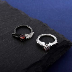 K&ouml;nnte beinhalten: Zwei dekorative Ringe mit herzf&ouml;rmigen roten Edelsteinen. Ein Ring ist schwarz mit Fl&uuml;gelakzenten, der andere silberfarben. Die Ringe werden auf einer dunklen, strukturierten Oberfl&auml;che vor blauem Hintergrund pr&auml;sentiert.