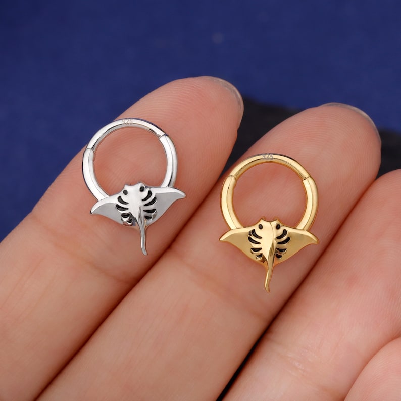 May include: Two hoop earrings, one silver and one gold, each featuring a manta ray design. The manta ray has a detailed, stylized appearance with curved lines and a pointed tail. The earrings are shown against a blue background.