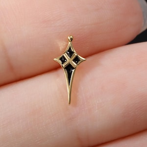 May include: A small, gold-toned metal charm shaped like a four-pointed star or cross with elongated, tapered ends. The centre features a recessed black enamel inlay with a raised gold-coloured cross symbol positioned directly in the middle.