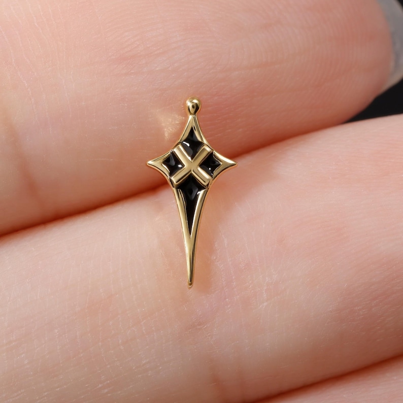 May include: A small, gold-toned metal charm shaped like a four-pointed star or cross with elongated, tapered ends. The centre features a recessed black enamel inlay with a raised gold-coloured cross symbol positioned directly in the middle.