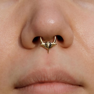 Puede incluir: Un anillo de tabique dorado con un diseño de manta raya. La joya se lleva en la nariz, con el colgante de manta raya centrado debajo de las fosas nasales. El anillo tiene un acabado pulido y está sobre un fondo de piel.