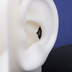 May include: A silver ear piercing stud with a black crescent moon design, sun, and starburst detail. The jewellery is set against a white ear model, with a dark blue background.