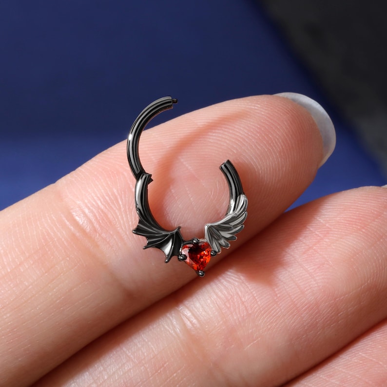 K&ouml;nnte beinhalten: Schwarzer Septumring mit einem herzf&ouml;rmigen roten Edelstein. Der Ring hat Fledermausfl&uuml;gel und Engelsfl&uuml;gel auf beiden Seiten des Edelsteins. Der Schmuck ist aus dunklem Metall gefertigt und f&uuml;r das Tragen in der Nase konzipiert.