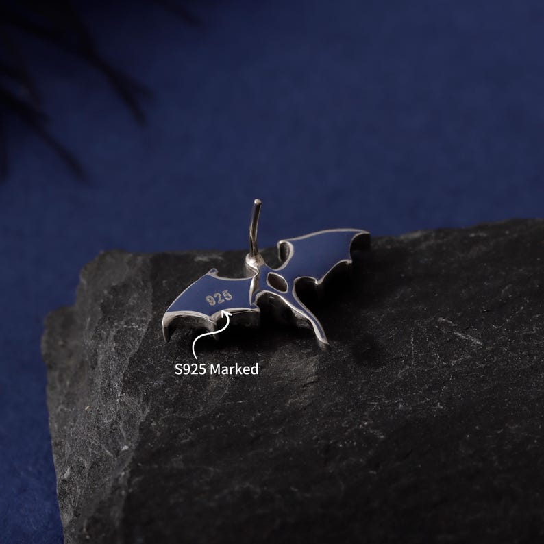 May include: A silver-colored earring with a blue enamel bat wing design. The earring is marked with "925" and "S925 Marked". The bat wings are spread out, and the earring has a post for piercing.