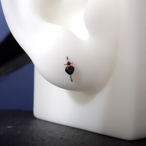 May include: A silver earring featuring a black heart-shaped gem and a small red gem. The earring is designed with a vertical bar and a decorative top, showcasing a gothic or romantic aesthetic.
