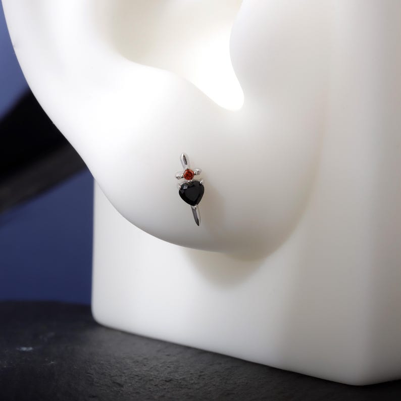 May include: A silver earring featuring a black heart-shaped gem and a small red gem. The earring is designed with a vertical bar and a decorative top, showcasing a gothic or romantic aesthetic.