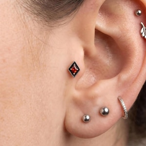 Könnte beinhalten: Ein rautenförmiger Tragus-Ohrstecker mit einem zentralen roten Edelstein in einer dunklen Metallfassung mit Perlenrand. Das Ohr zeigt zudem zwei silberne Kugelstecker, einen kleinen silbernen Ring mit klaren Steinen und einen blattförmigen Knorpelstecker.
