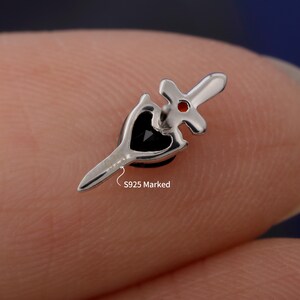 May include: A silver-colored earring with a dagger design, featuring a black heart-shaped stone and a red accent. The earring is marked "S925".