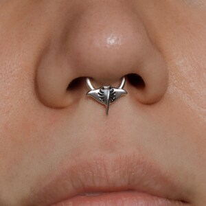 May include: A silver septum ring featuring a manta ray design. The jewelry is centered on the nose, with the manta ray pendant hanging below the septum. The ring has a polished finish and detailed etching.