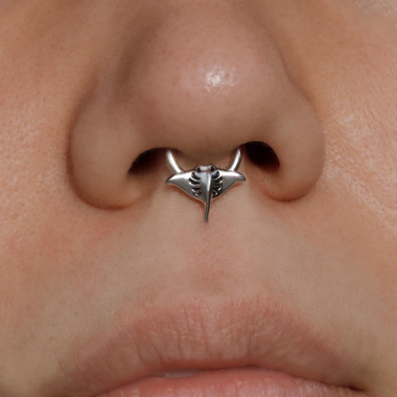 May include: A silver septum ring featuring a manta ray design. The jewelry is centered on the nose, with the manta ray pendant hanging below the septum. The ring has a polished finish and detailed etching.