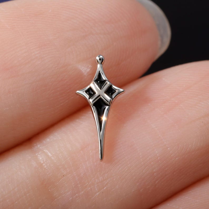 May include: A small, silver-toned metal charm shaped like a four-pointed star or cross. The design features a polished metallic finish with black enamel inlay filling the recessed areas between the central cross shape and the outer pointed edges.