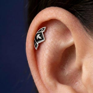 Op de afbeelding: Een zilveren oorbel met een zwart-wit bladontwerp. De sieraden zijn geplaatst tegen de achtergrond van een oor, waarbij de ingewikkelde details van de piercing worden getoond. Het ontwerp heeft een gestileerd bladmotief.