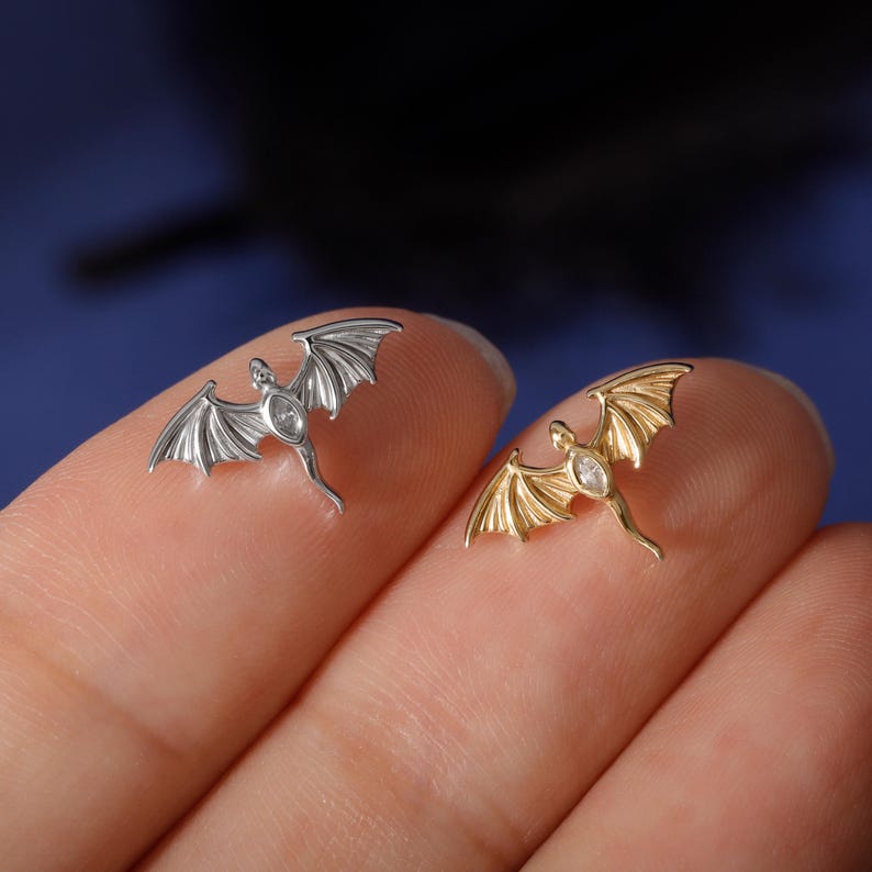 May include: Two small dragon-shaped pendants, one silver and one gold, each with a clear gemstone in the center. The pendants have detailed wing designs and are held between fingers against a blue background.