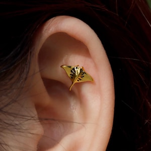 Peut inclure: Boucle d'oreille dorée en forme de raie manta, ornée de détails noirs sur les ailes et le corps. Ce petit bijou délicat est porté sur une oreille, ajoutant une touche originale.