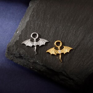 May include: Two dragon-shaped pendants, one silver and one gold, are shown on a dark, textured surface. Each pendant has outstretched wings, a tail, and a small gemstone. The pendants are designed for jewelry.