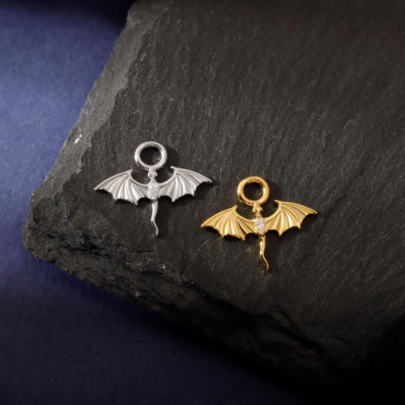 May include: Two dragon-shaped pendants, one silver and one gold, are shown on a dark, textured surface. Each pendant has outstretched wings, a tail, and a small gemstone. The pendants are designed for jewelry.