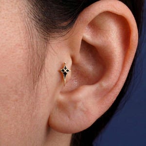 Könnte beinhalten: Ein goldfarbener Ohrstecker in Form eines vierzackigen Sterns mit schwarzer Einlage. Das Schmuckstück ist am Tragus des Ohrs platziert und zeigt ein symmetrisches Design mit scharfen, länglichen Spitzen und einem zentralen Kreuzdetail.
