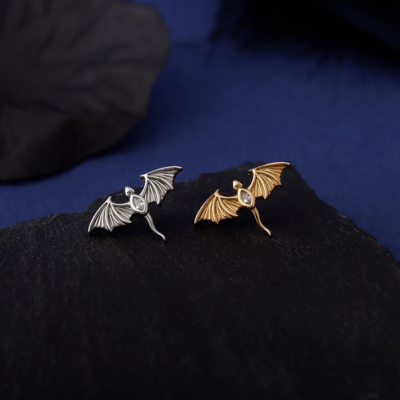 May include: Two dragon-shaped brooches, one silver and one gold, each with a small, clear gemstone. The dragons have outstretched wings and detailed wing patterns. They are displayed on a dark, textured surface against a dark blue background.