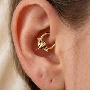 May include: A gold-colored daith piercing hoop with a detailed manta ray charm. The charm has intricate cut-out designs and is positioned within the ear's cartilage. The jewelry is shown in a close-up shot.