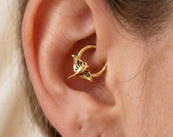 May include: A gold-colored daith piercing hoop with a detailed manta ray charm. The charm has intricate cut-out designs and is positioned within the ear's cartilage. The jewelry is shown in a close-up shot.